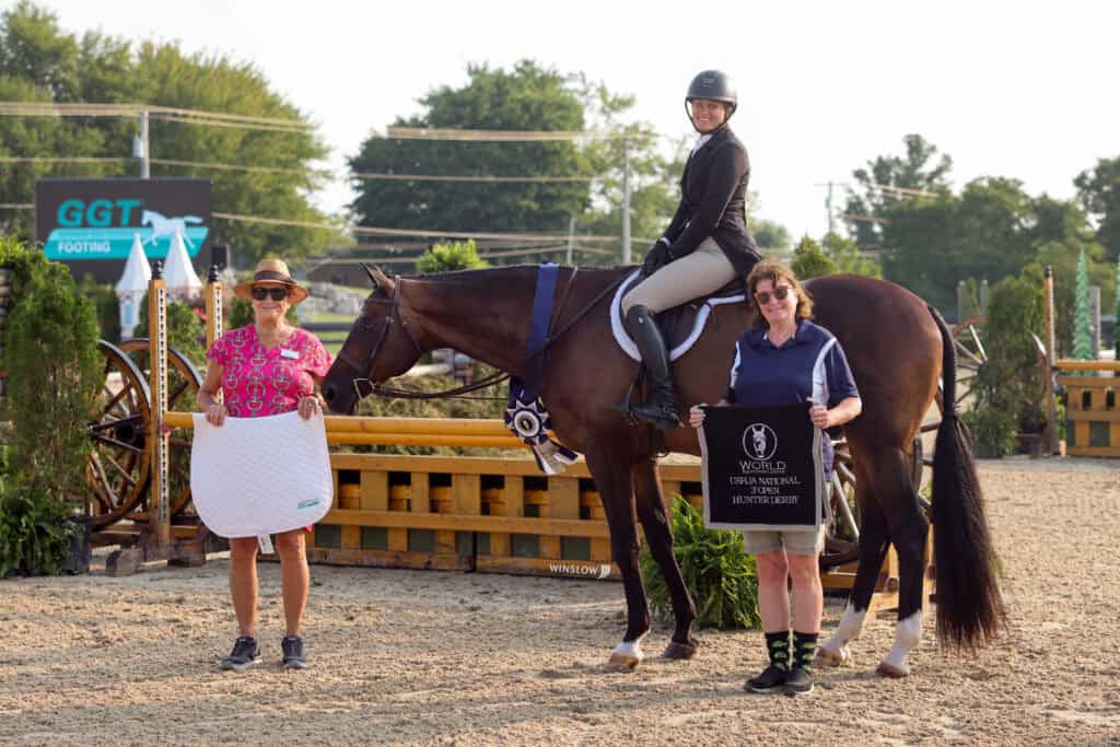 Three people stand with a horse at an outdoor equestrian event; one person is mounted, while the other two hold award items and smile for the camera.