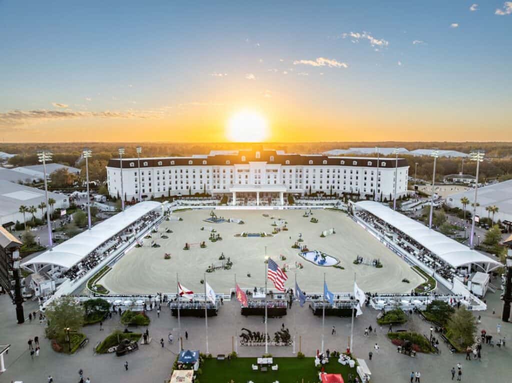 A large equestrian arena with jumps is set up in front of a grand hotel at sunset, with flags and spectators visible in the foreground.