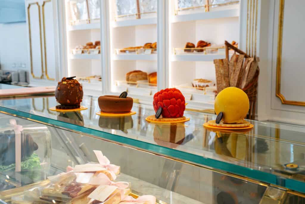 Four assorted gourmet pastries are displayed on a glass counter in a bakery, with shelves of bread and baguettes in the background.