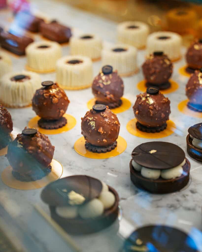 A display of assorted gourmet desserts, including chocolate-covered spheres, layered pastries, and molded white confections, arranged on a marble surface.
