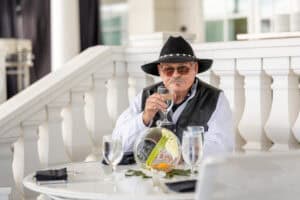An older man in a black hat and vest sits at an outdoor table holding a glass of water, with three more glasses and a small floral centerpiece in front of him.