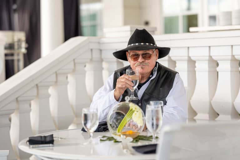An older man in a black hat and vest sits at an outdoor table holding a glass of water, with three more glasses and a small floral centerpiece in front of him.