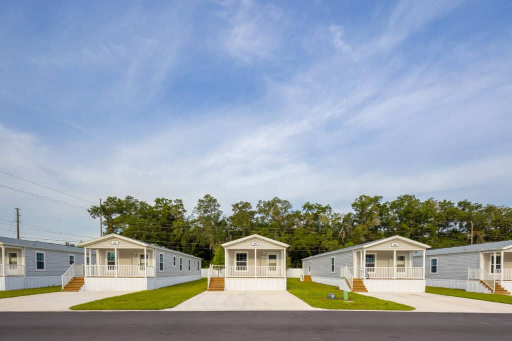 Three identical manufactured homes with driveways are lined up on a neat, grassy lot under a blue sky, with trees and power lines in the background.