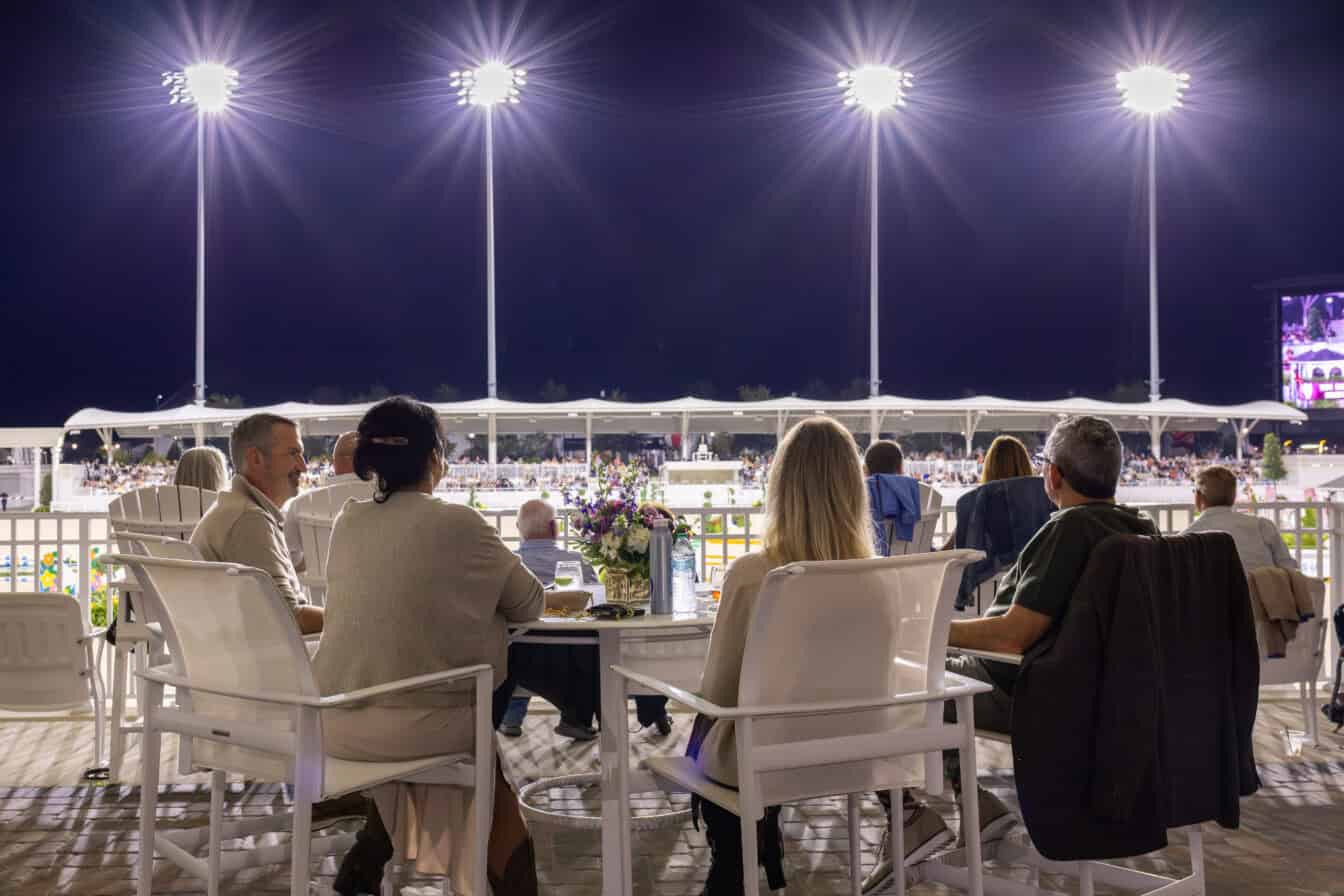 People seated at outdoor tables watch a nighttime sporting event under bright stadium lights, with a flower arrangement on one table and a large screen visible in the background.