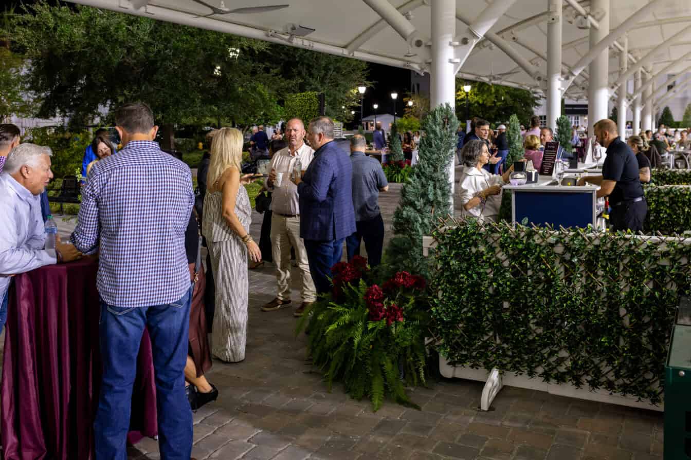 People socialize and enjoy drinks at an outdoor evening event under a canopy, surrounded by greenery and festive decorations.