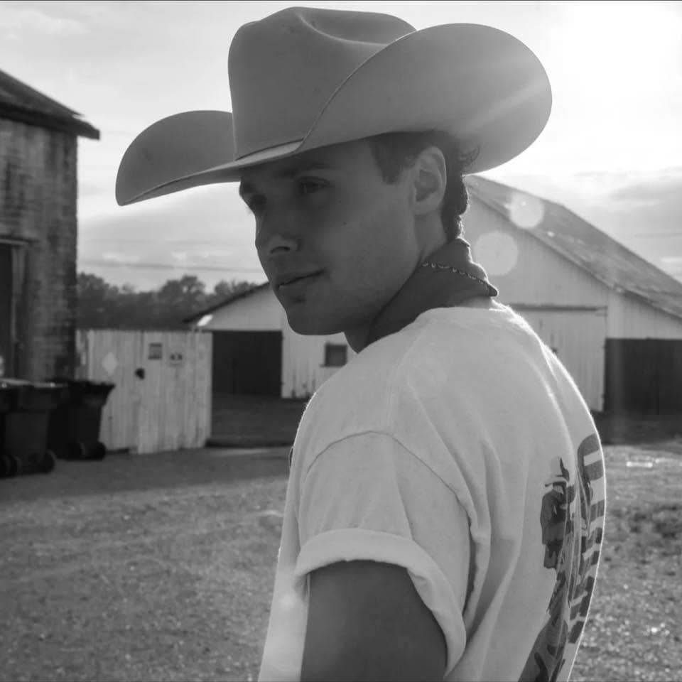 A person wearing a cowboy hat and t-shirt stands outside near barns and sheds on a gravel lot, looking over their shoulder.