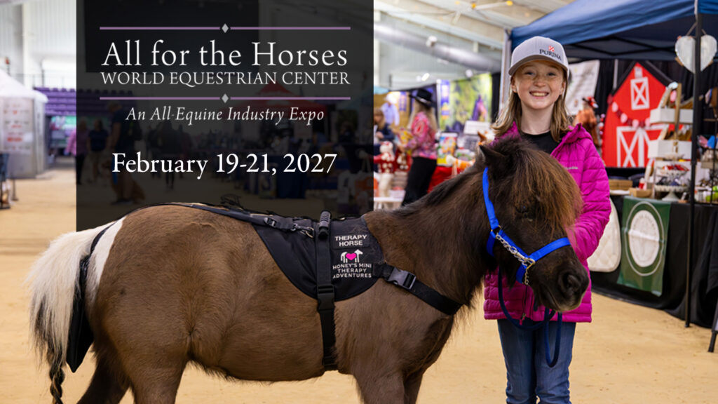 A girl in a pink jacket smiles and stands beside a small therapy horse at an indoor equestrian expo with vendor booths in the background.
