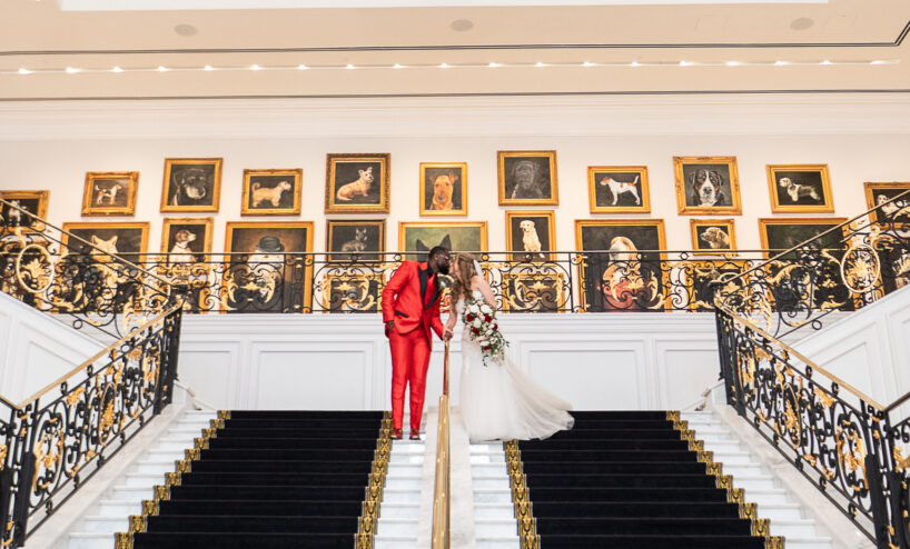 A couple in formal attire stands together at the center of a grand staircase, surrounded by ornate railings and framed portraits on the wall behind them.