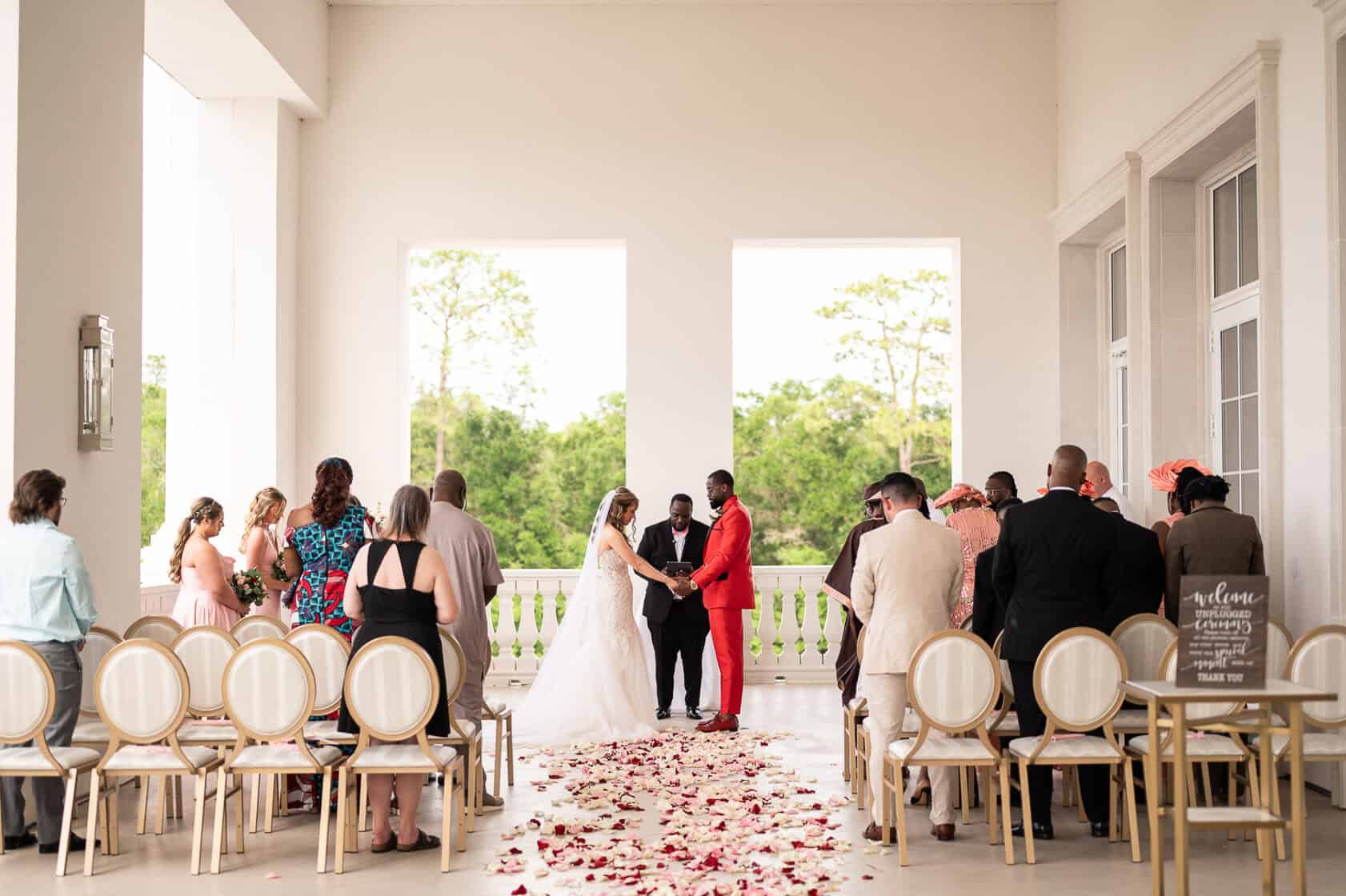 A bride and groom stand facing each other at an indoor wedding ceremony, holding hands, with guests seated and flower petals scattered on the floor.