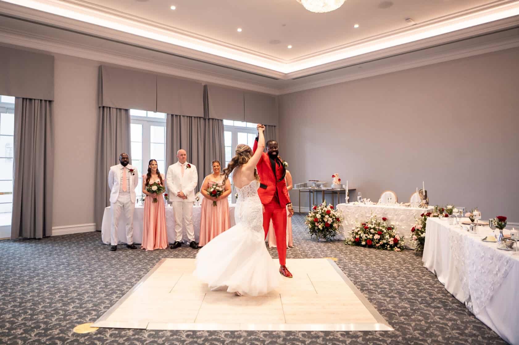 A bride and groom dance in the center of a decorated ballroom, with bridal party members in the background watching and smiling.