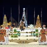 A rider and horse jump over an obstacle decorated with gingerbread figures at a nighttime equestrian event with lit Christmas trees in the background.
