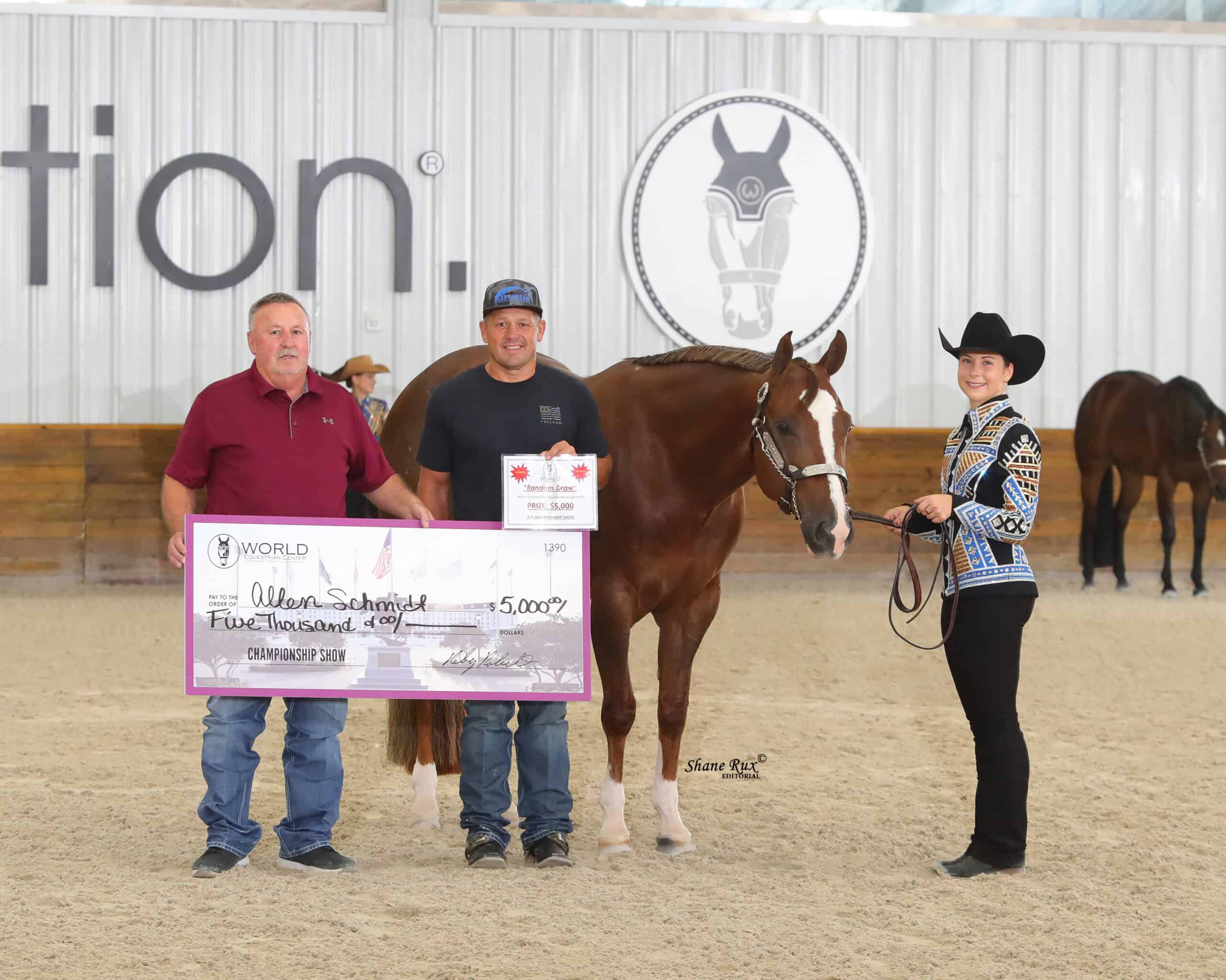 Three people and a horse stand indoors; one person holds a trophy, another holds a $5,000 check, and a woman in western attire stands beside the horse.