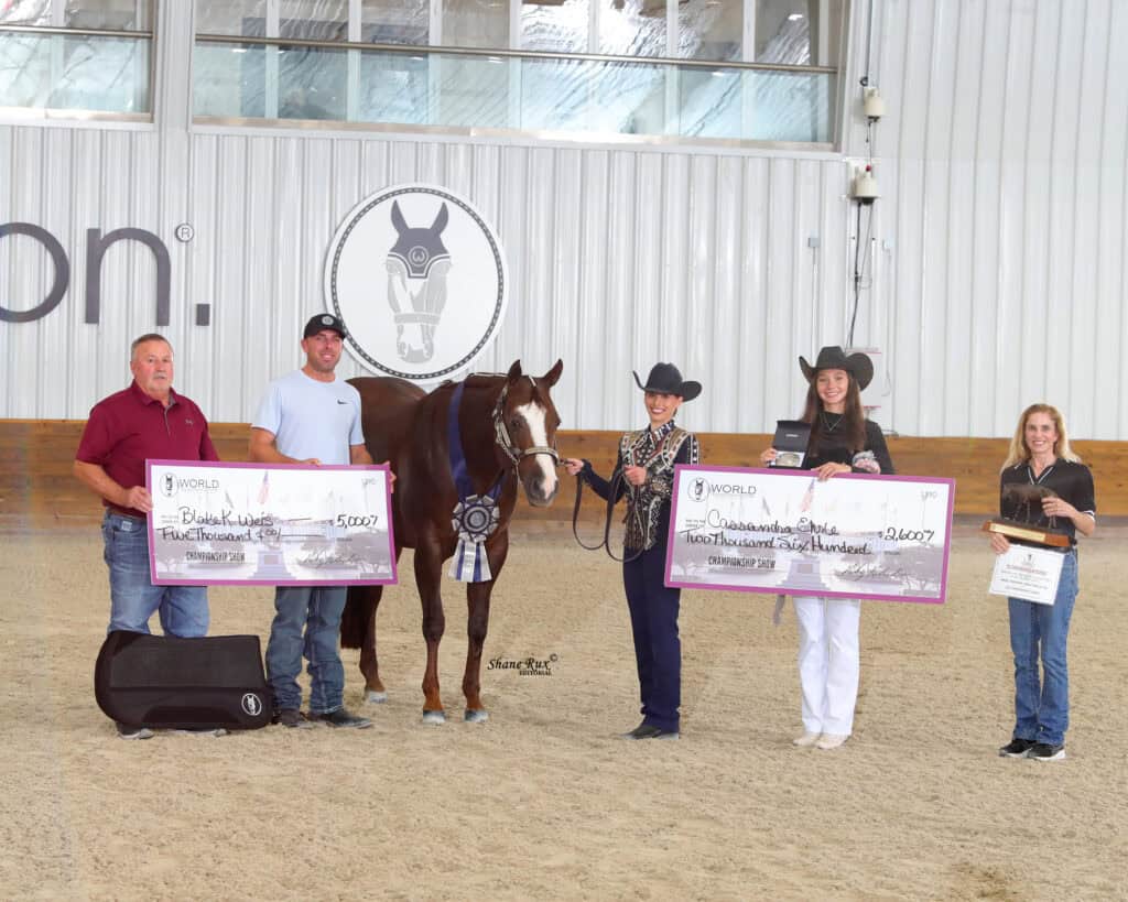 Five people and a horse stand indoors. Two women hold oversized checks, one man holds a case, and everyone is smiling. The horse wears a ribbon. An equestrian logo is on the back wall.