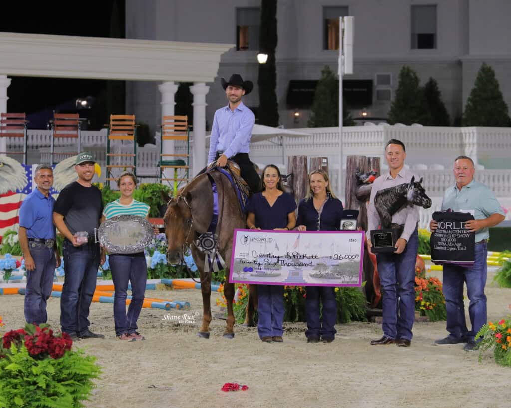 A group of people pose with prizes and a large check next to a man on horseback at an outdoor event with flowers and decorations.