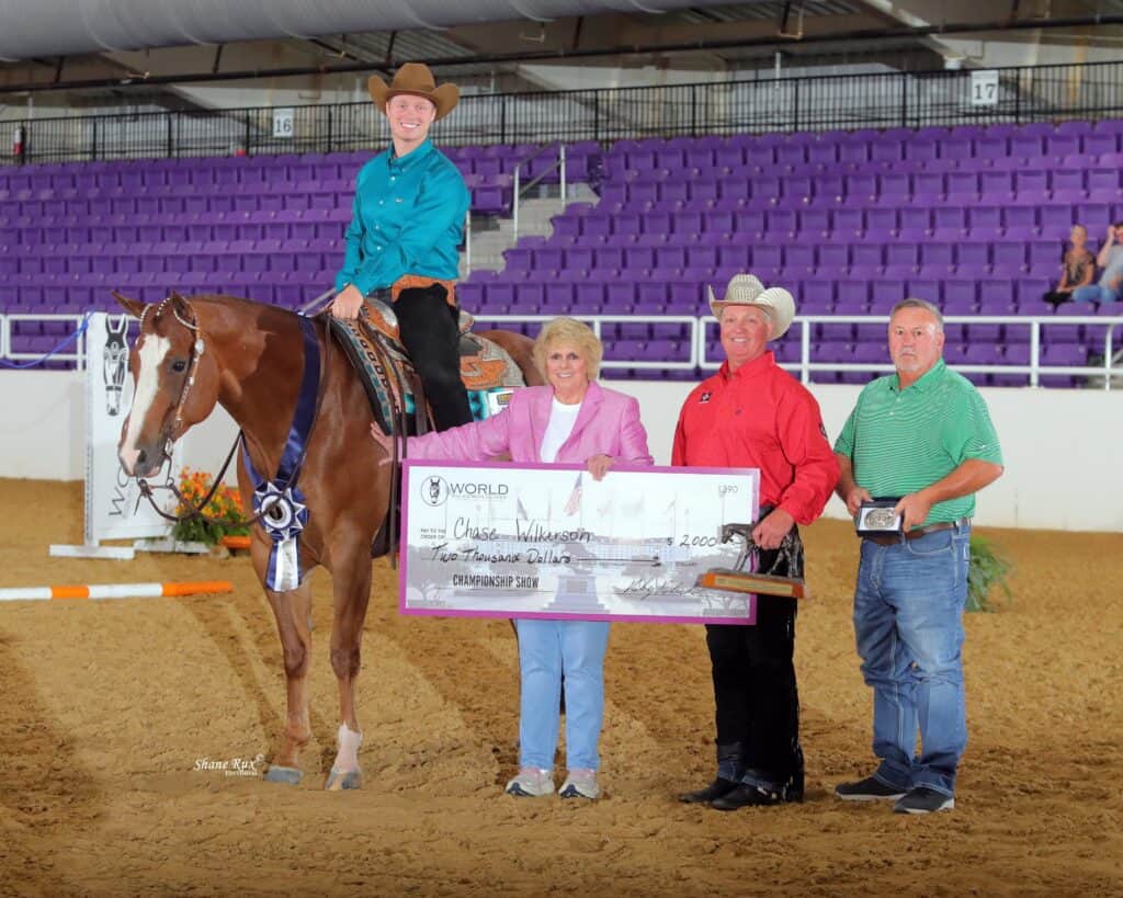 A person on horseback and three people standing present an oversized $2,000 check in an indoor arena with purple seats in the background.