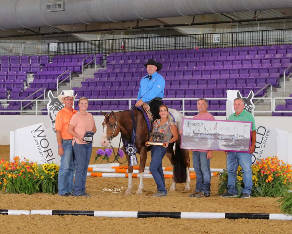 A group of six people pose with a horse in an indoor arena; one holds a giant check, another holds trophies, and the bleachers in the background are empty.
