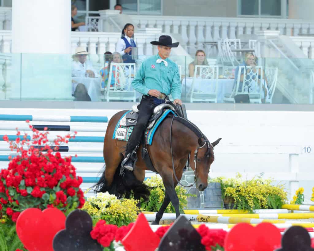 A man in a teal shirt and black hat rides a brown horse through an arena with flowers and colorful poles, as spectators watch from the stands.