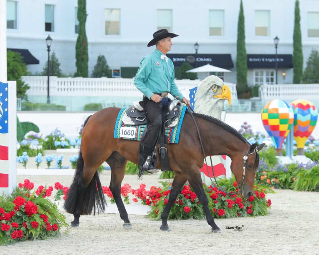 A man in a teal shirt and black hat rides a brown horse, numbered 1660, in an arena decorated with flowers and patriotic props, including a large eagle head.