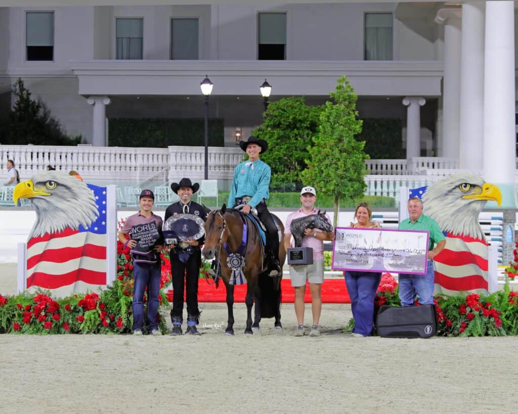 A group of six people and a horse pose in front of patriotic eagle decorations, holding awards, flowers, and a large check in an outdoor arena.