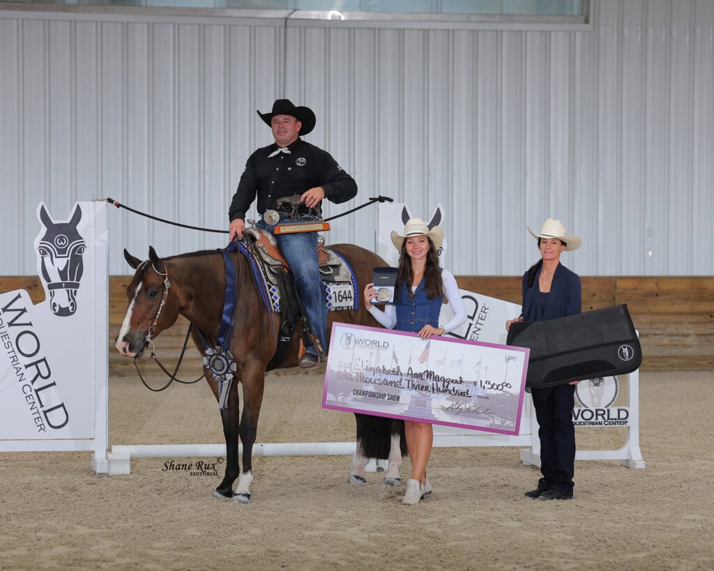A person on horseback poses with two people standing, one holding a large check and trophy. All are dressed in western attire inside an indoor arena.