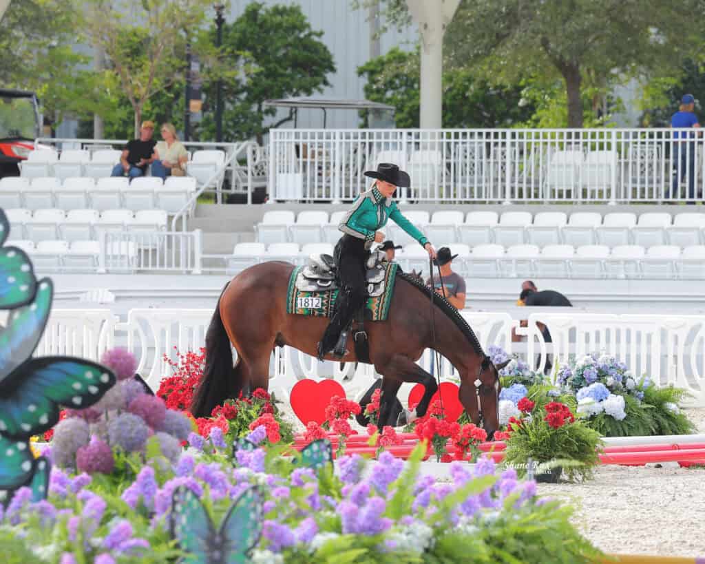 A rider in western attire guides a brown horse over red poles surrounded by colorful flowers in an outdoor equestrian arena with empty white seats in the background.