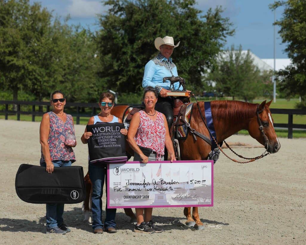 Four women and one person on horseback pose outdoors holding a large check for $5,200, a trophy, and a banner. Trees and a fence are visible in the background.