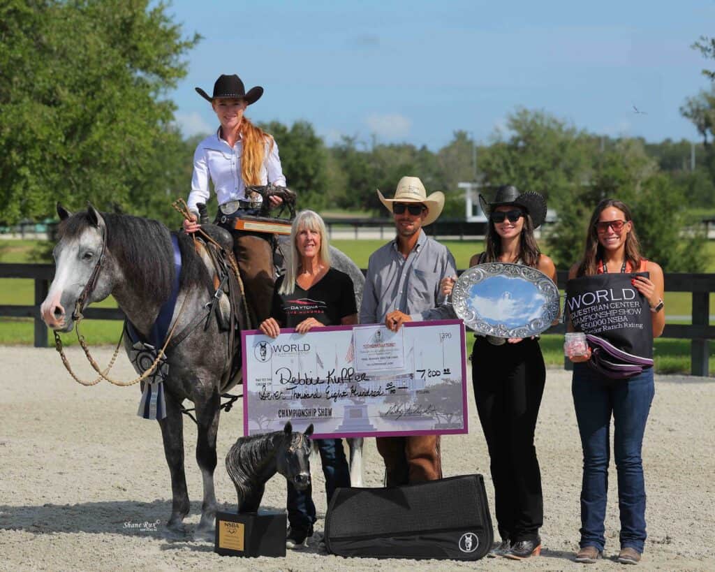 A woman on a horse poses with four people holding a large check, a silver plate, and a plaque at an outdoor equestrian event.
