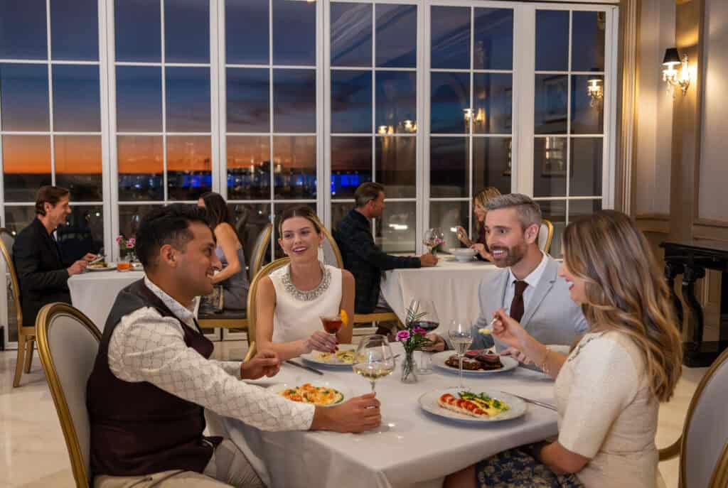 Four people dine together at a restaurant table, smiling and conversing, with other diners in the background and a sunset visible through large windows.