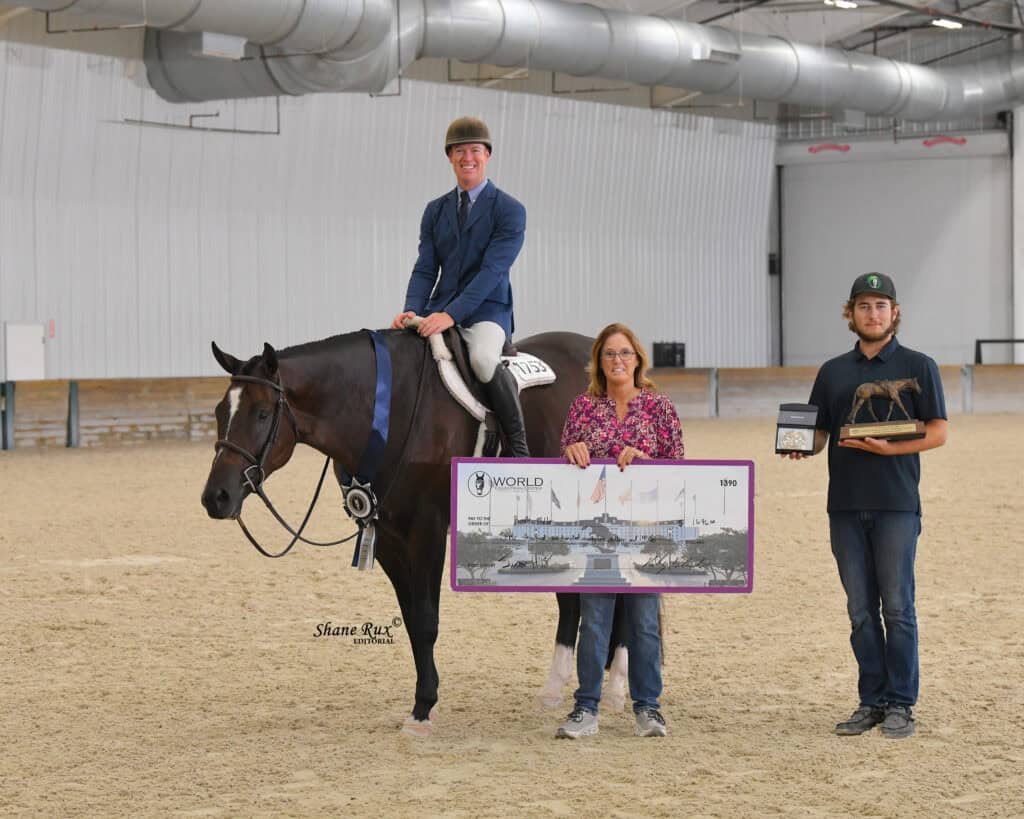 A person on horseback poses indoors with two people standing, all holding a large World Championship sign, in an equestrian arena.