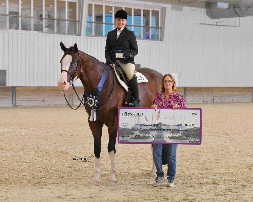 A rider on a horse stands beside a woman holding a large award sign in an indoor arena with spectator seating in the background.