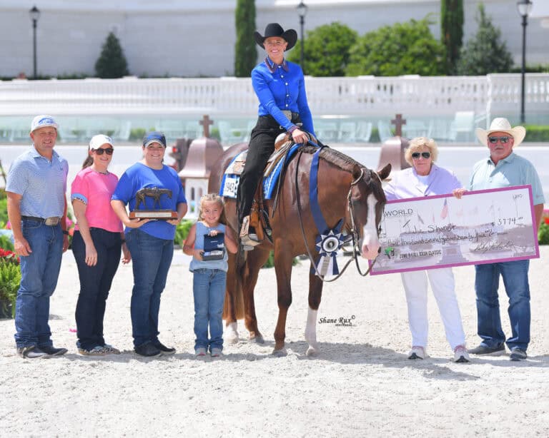 A group of people stands next to a woman on a horse. The group holds a trophy and a large check. The scene is outdoors in an arena.