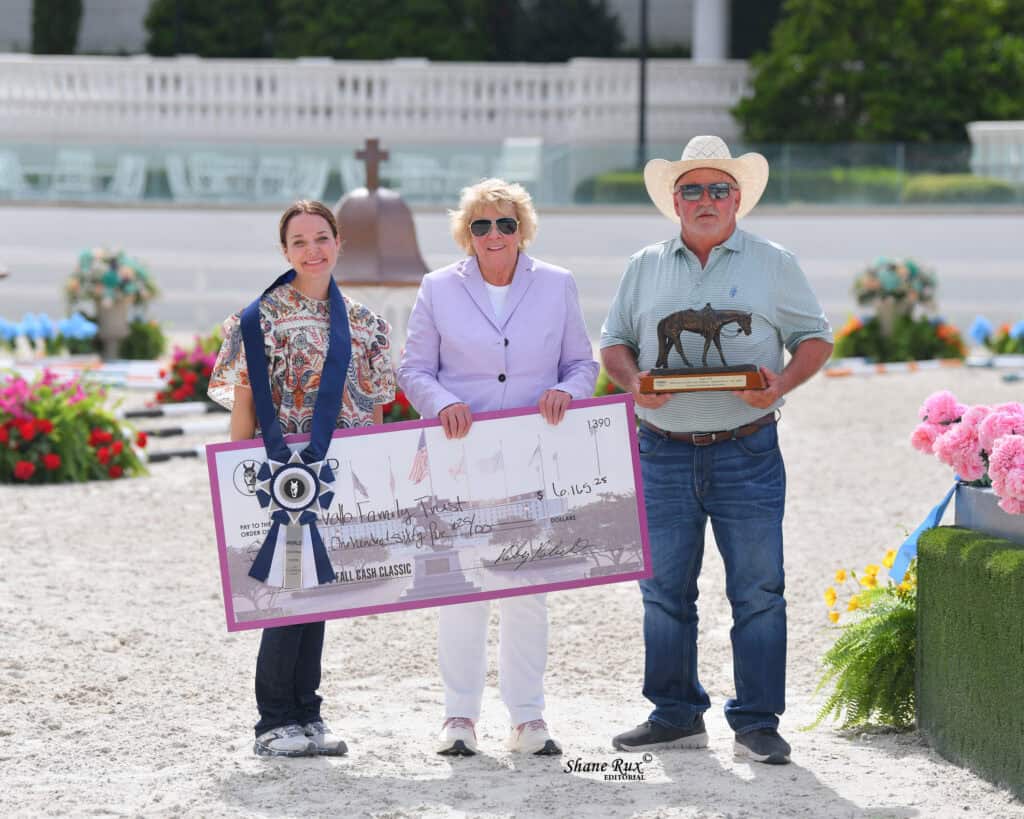 Three people stand outside holding a large ceremonial check, a trophy, and wearing medals, with flowers and equestrian jumps visible in the background.