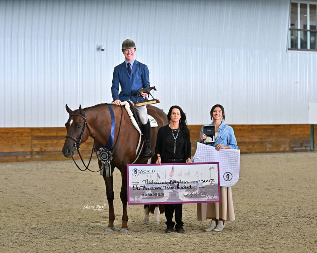 A person on horseback poses indoors beside two people standing, one holding a large check and the other holding a white mat. All are smiling and dressed formally.