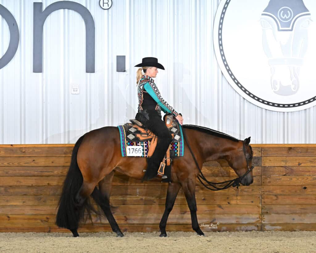 A rider in a black hat and patterned shirt rides a brown horse with decorative tack inside an indoor arena with wooden walls and a large logo on the wall.