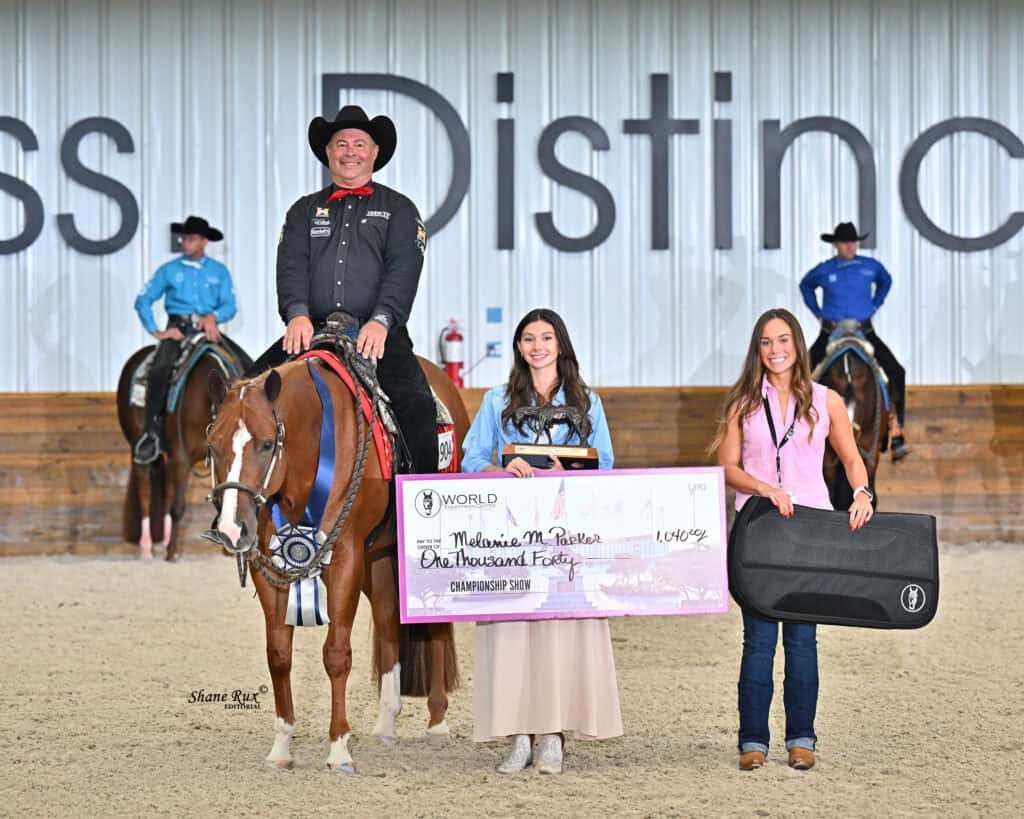 A man on horseback and two women stand indoors, one holding a large check and the other a case. Three people on horses are in the background, with a partially visible event banner behind them.