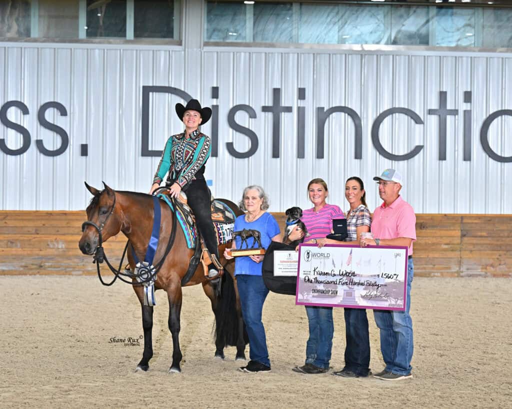 A woman on a horse poses with four people holding a trophy and an oversized check in an indoor arena.