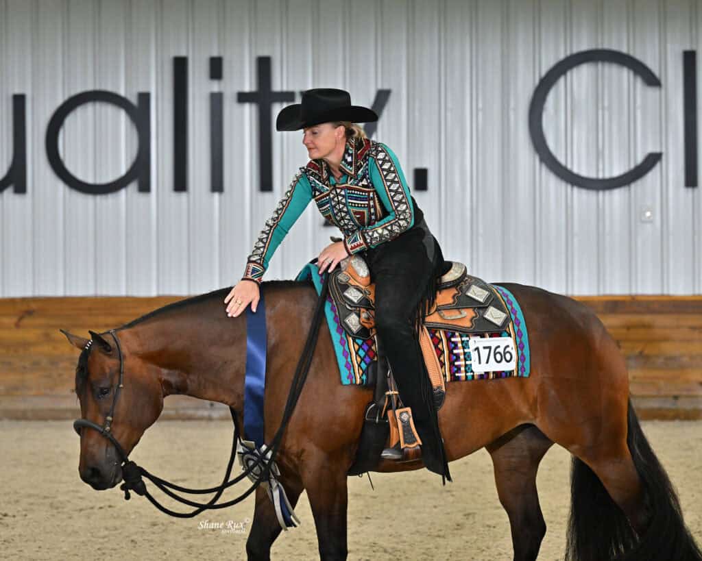 A woman in a patterned outfit and black hat touches her brown horse’s neck, holding a blue ribbon, inside an indoor arena.