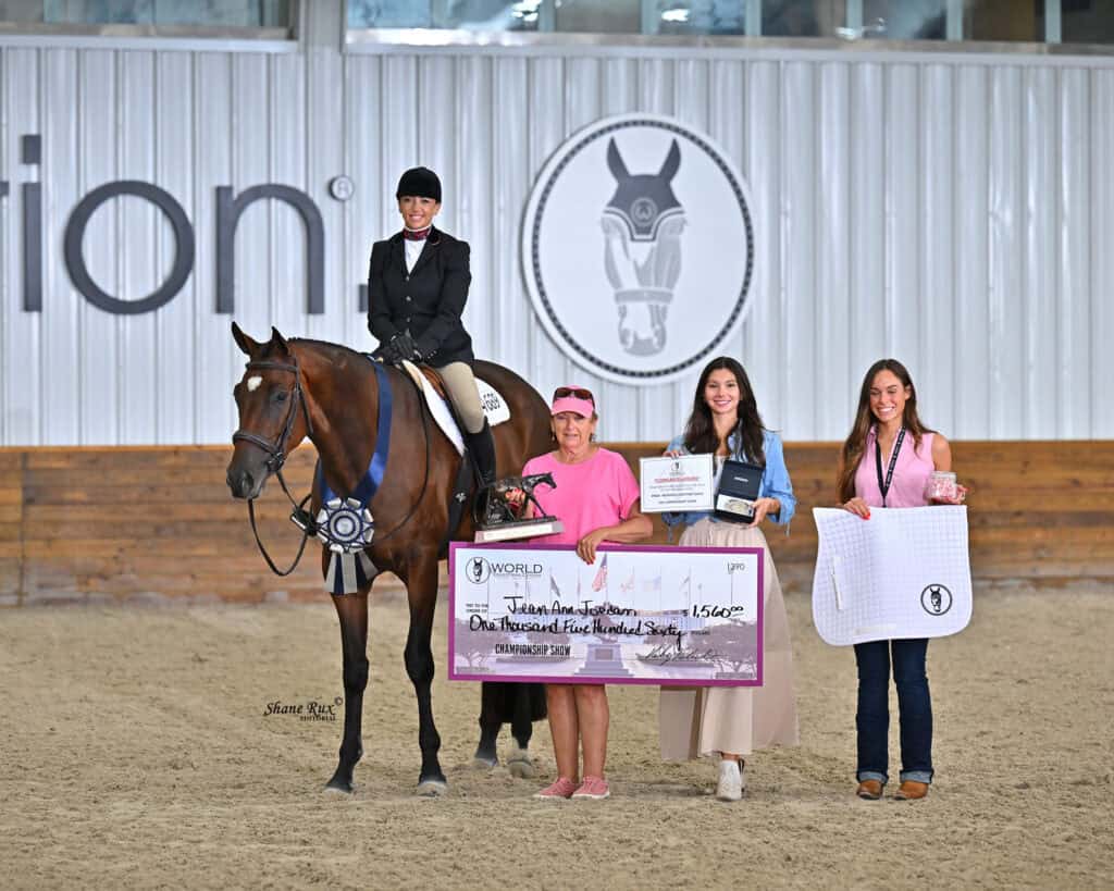A person on horseback with a blue ribbon poses beside three people holding a large check, a plaque, and a white saddle pad in an indoor equestrian arena.