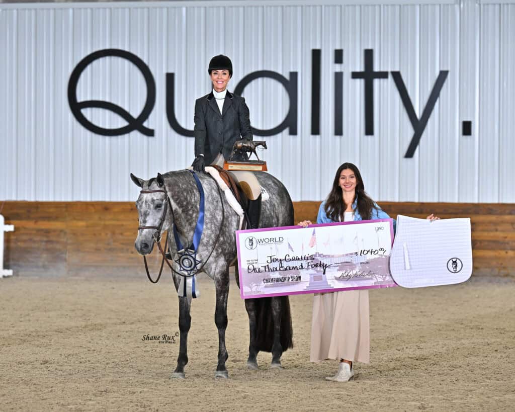 A person on horseback and another standing hold a large award check and a ribbon in an indoor arena with “Quality” on the wall behind them.
