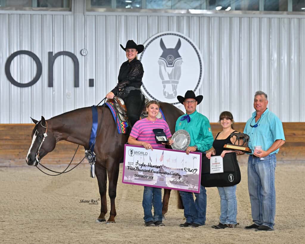 A rider on a horse poses with five people holding a large check, trophies, and awards in an indoor arena.