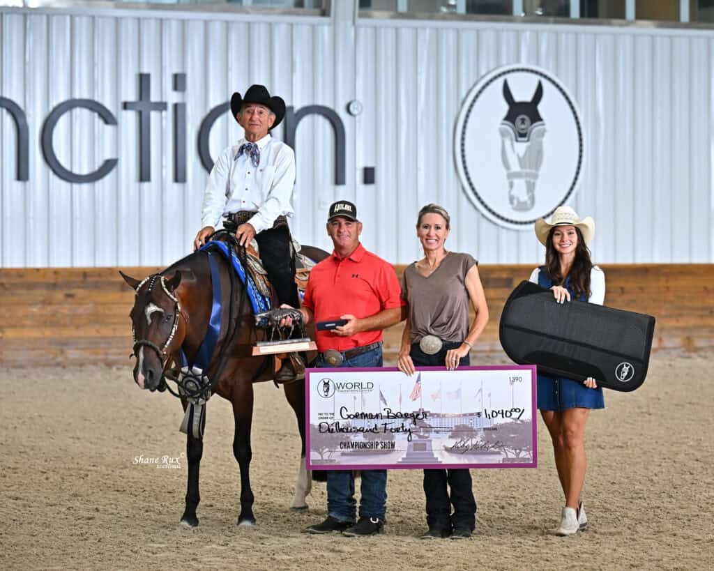 Four people pose with a horse in an indoor arena, holding a large check and an award case. The rider wears a suit and cowboy hat. A painted horse and “World” text are visible in the background.
