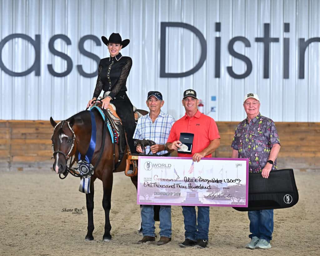A woman on a horse poses with three men indoors, holding a large check for $1,300 and awards in front of a sign partially reading “ass Disti.”.