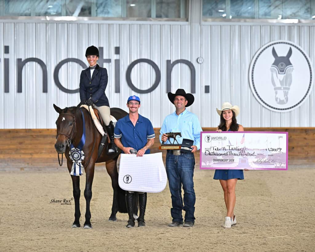 Four people and a horse pose indoors; one person is mounted, two stand with awards, and one holds a large check. A banner with a horse logo and partial text is visible in the background.