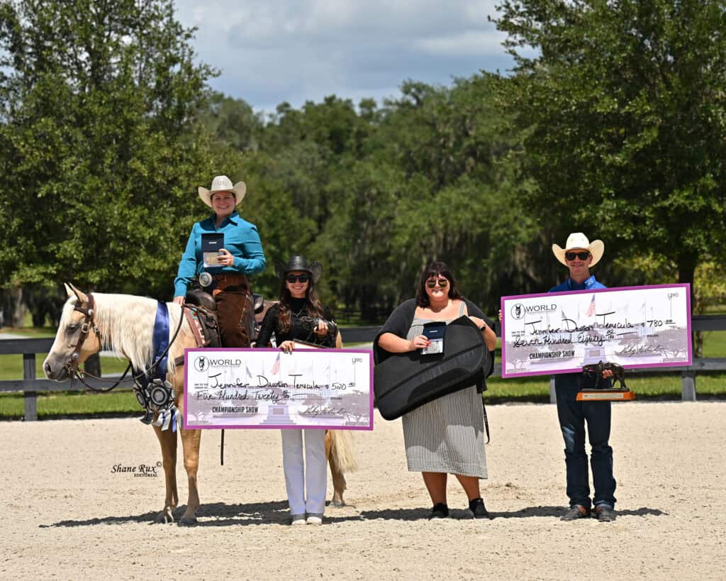 Four people pose outdoors, three holding large prize checks and trophies; one is seated on a horse. Trees and a fence are in the background under a partly cloudy sky.