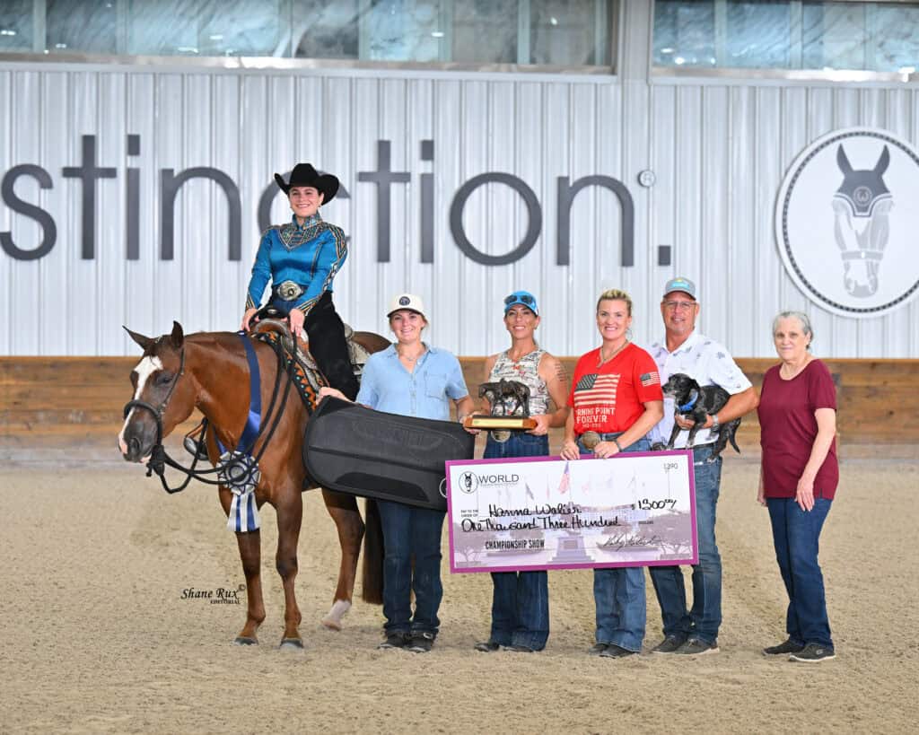A group stands in an arena; one person sits on a horse while others hold a large check, trophies, and a ribbon. A branded wall and dirt floor are visible in the background.