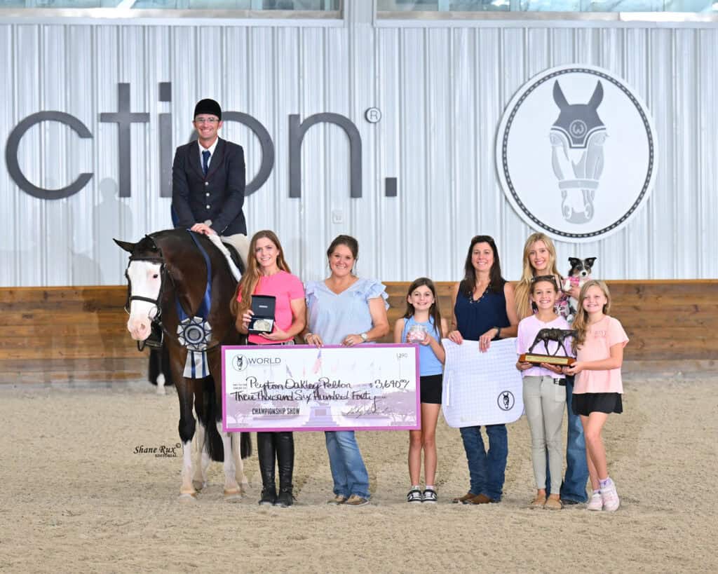 A group of people and a person on a horse pose indoors with a large check, trophy, and awards in an equestrian arena.