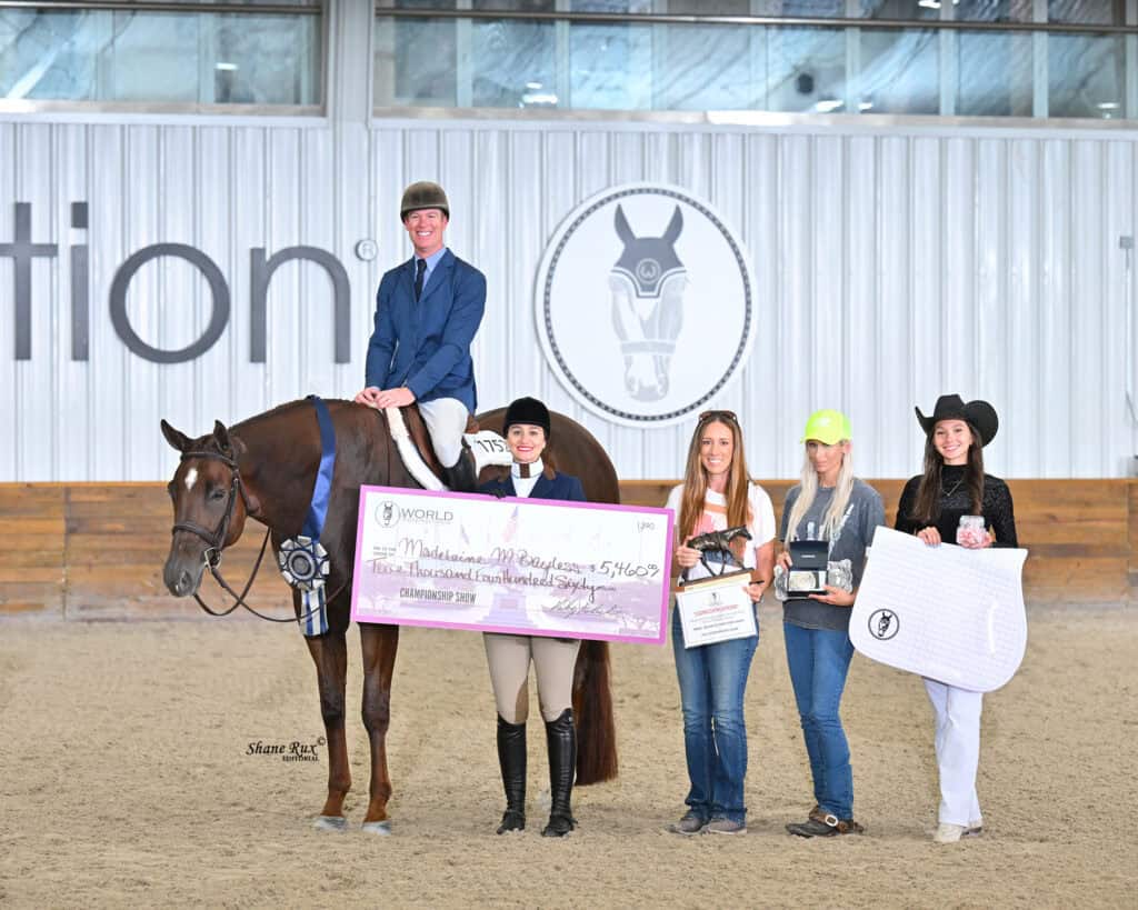 A rider on a horse poses with five people holding a large check and awards on an indoor arena with a horse logo on the wall behind them.