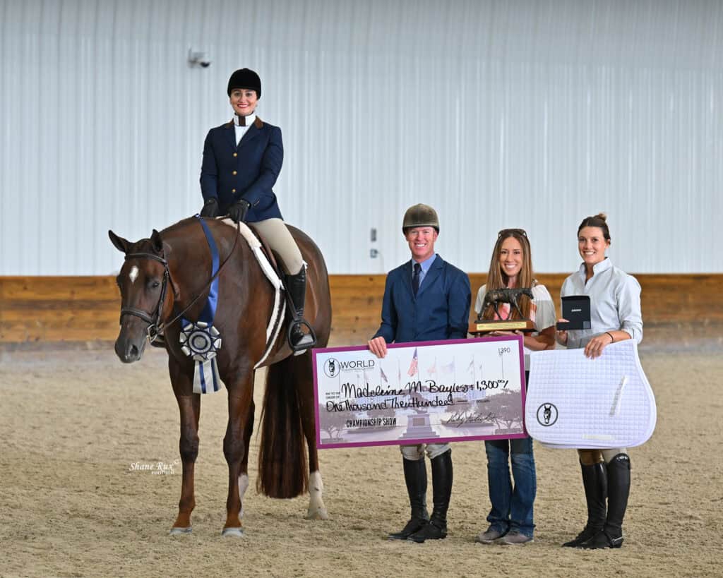 A rider on a horse poses with three people holding a large check, ribbons, and awards inside an indoor arena.