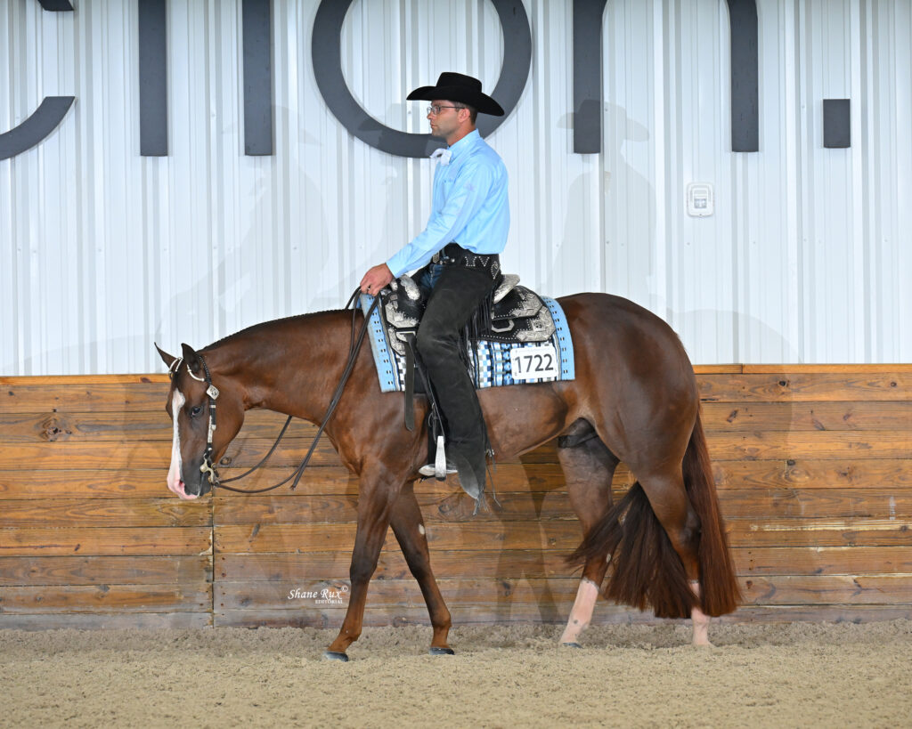 A man in a cowboy hat and blue shirt rides a brown horse with saddle number 1722 in an indoor arena with a wooden fence.