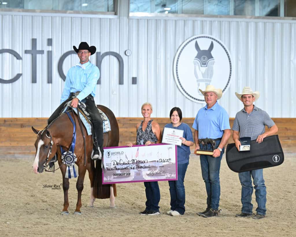 A man on horseback and four people stand indoors holding a large check, ribbon, and prizes at an equestrian event.